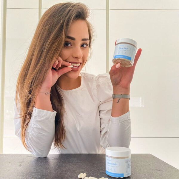 Woman with brown hair Holding Skinbiotics packaging in one hand and taking a capsule with the other, on table Skinbiotics packaging with capsules beside it